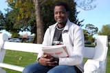 A man of African appearance sits on a white bench near a tree in a park. He is holding university textbooks.