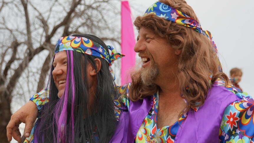 Two men, both dressed in colour clothes and bandanas and one wearing a wig, laugh.