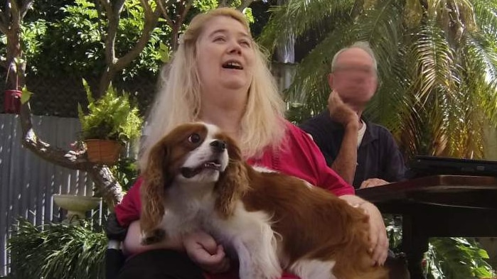 A woman sitting on a walking frame holding a small dog while a large dogs stands on grass.