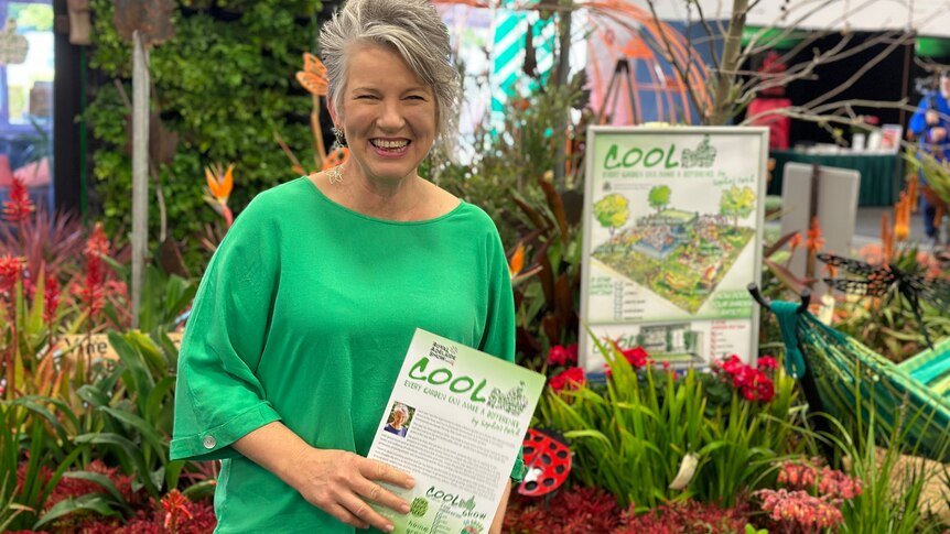 woman with grey hair in green top holding leaflet, with sign and plants in the background