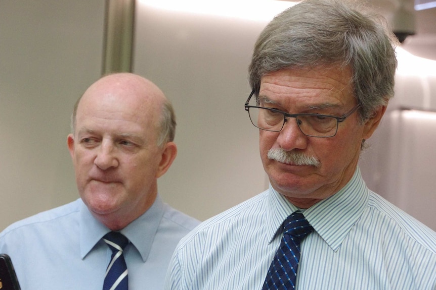 Mike Nahan looks down with John Day in the background at a media conference.
