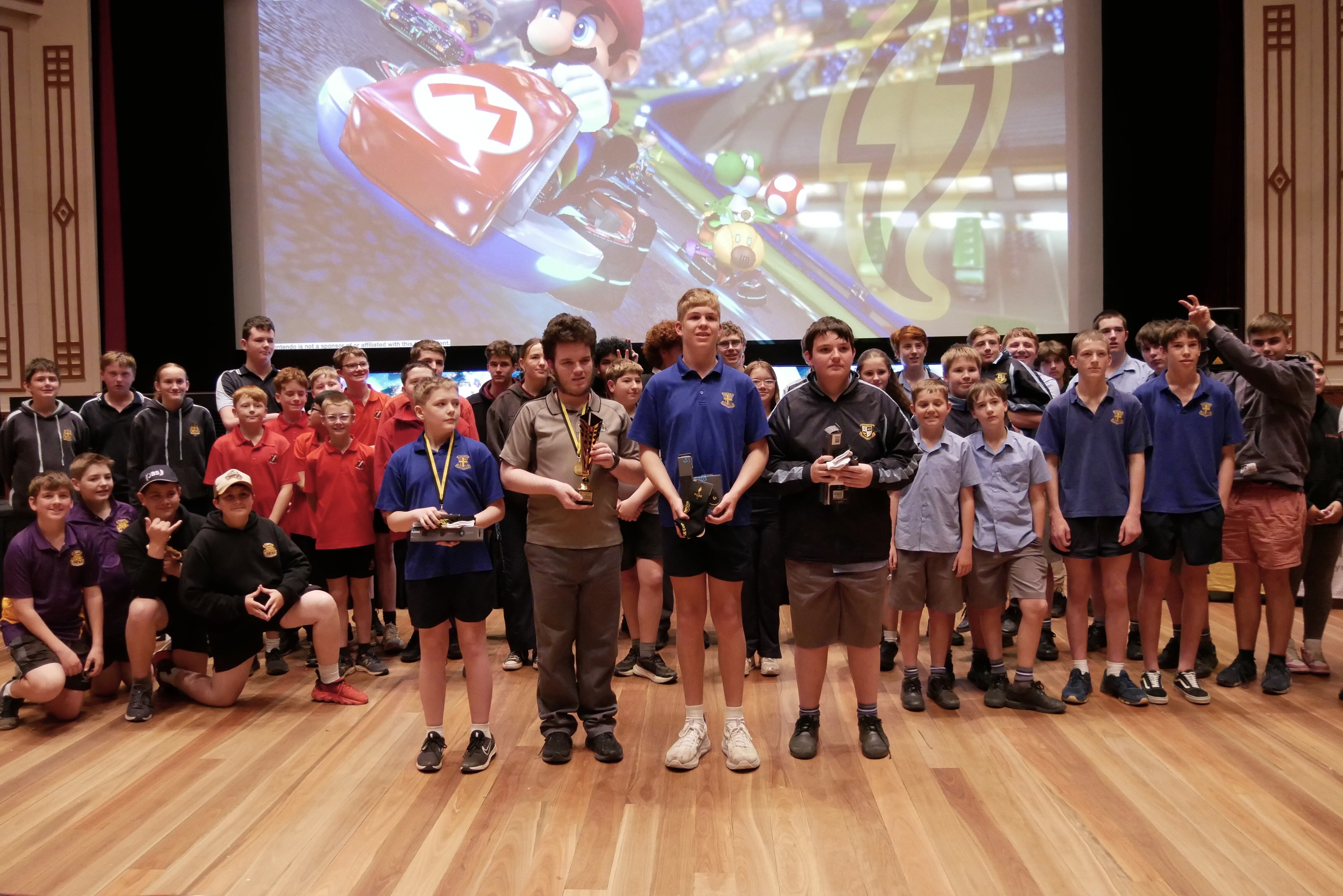 Four boys stand in front of a crowd of kids holding medals and trophies. 
