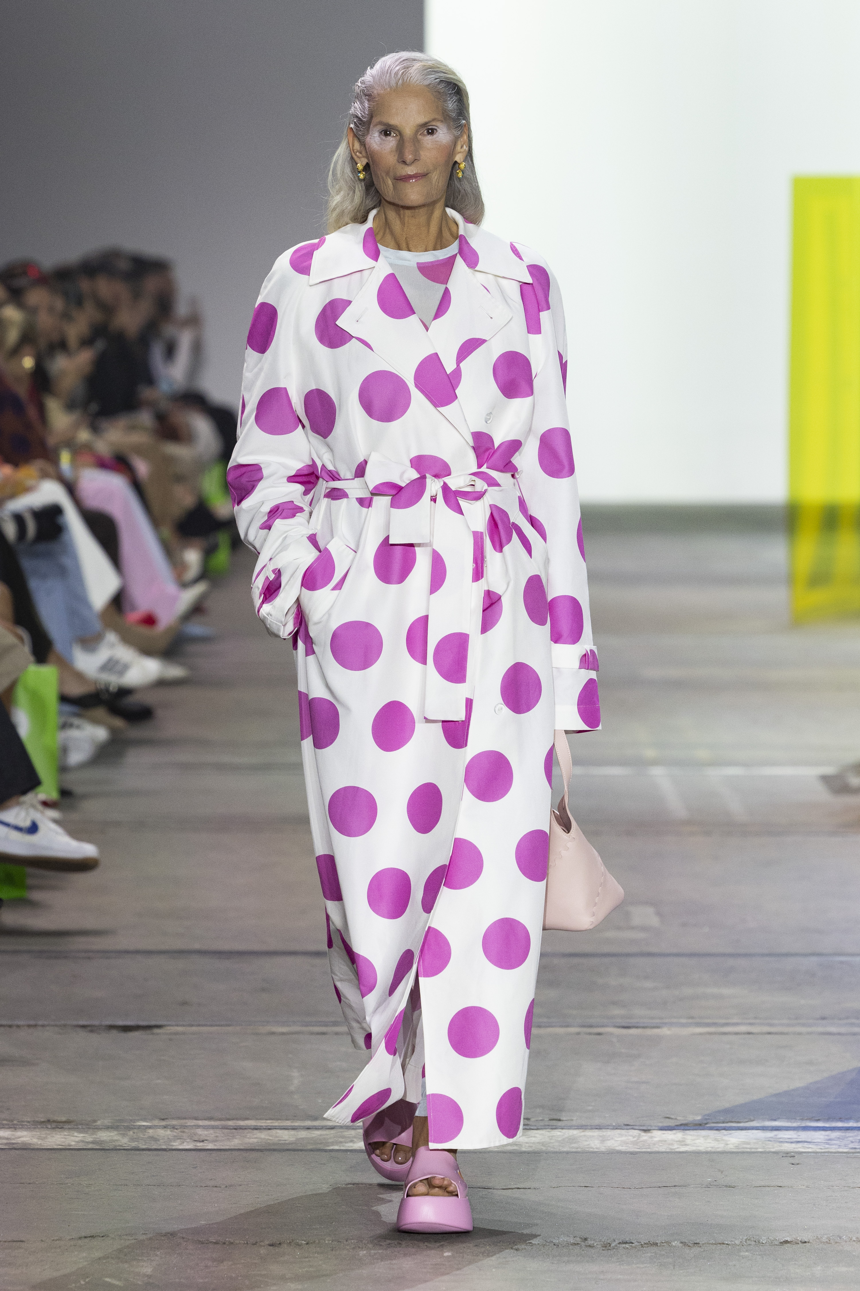 An elderly white woman with white hair models a white and pink polka-dot jumpsuit on a fashion runway.