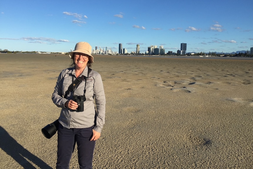 A woman wearing a hat and binoculars smiles while standing on a vast sandy bank with the Gold Coast skyline far in the distance