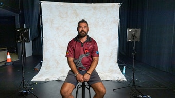 A man wearing a deep red top and dark grey shorts sitting on a stool on a TV set.