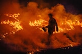 A firefighter with a can conducts backburning- he is a silhouette as a bushfire burns behind him.