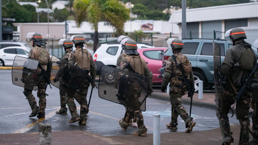 Troops in uniform walk away from the camera holding riot shields in the street