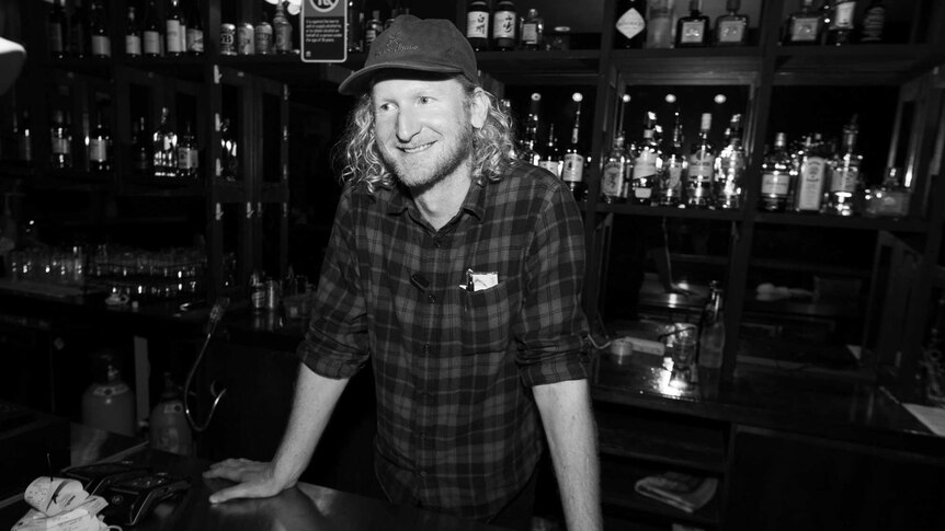 A black and white photo of a man standing behind a bar looking passed the camera and smiling.