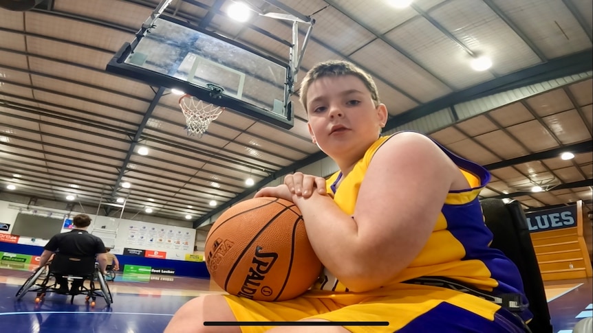A young boy sitting in a wheelchair wearing a yellow and purple singlet and shorts with a basketball on his lap.