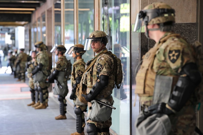A line of men dressed in camouflage uniforms and helmets stand outside an unmarked building.