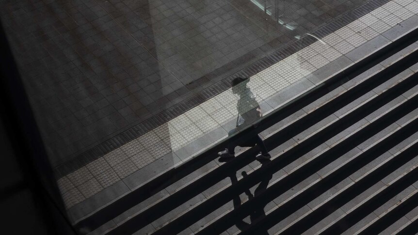 A young woman walks across steps at a university in Australia, half obscured by glass