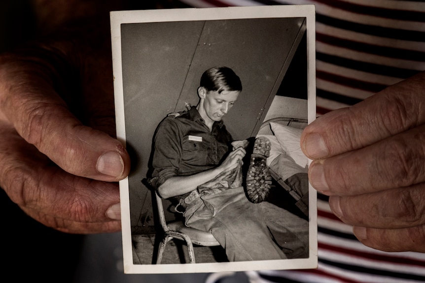 Hands hold a black and white photo of a young soldier cleaning boots.