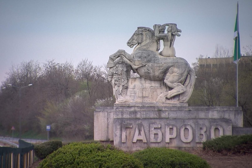 A stone horse statue with a person riding it is seen with Bulgarian writing on it, and flag to the rear. The sky is grey behind.