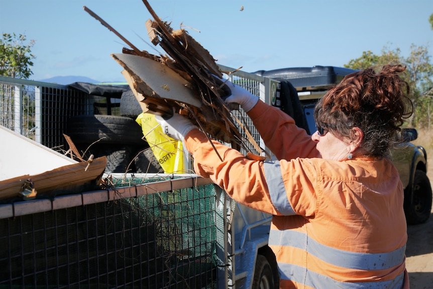 A middle aged woman with red hair and a high visibility shirt holds rotten chipboard above her head over a trailer. 