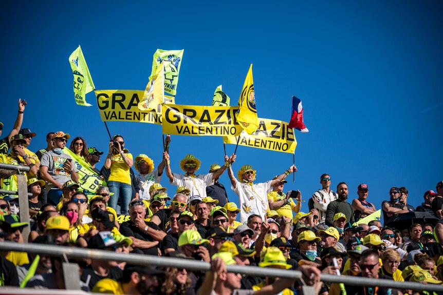 A section of fans in a grandstand, wearing some yellow clothing, hold signs saying 'Grazie Valentino' at a MotoGP race.