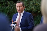 Mark McGowan wears a blue suit and pale pink tie, standing at microphones speaking to the media.