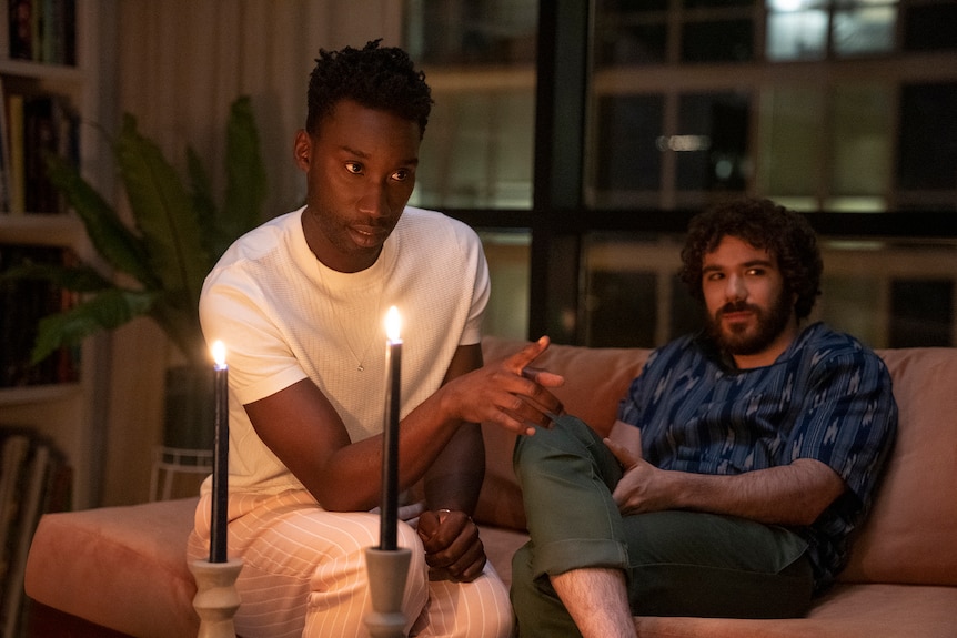 Troy Cartwright points a finger forewarningly in front of two lit candles while Grady Greenberg reclines with a look of reserve.