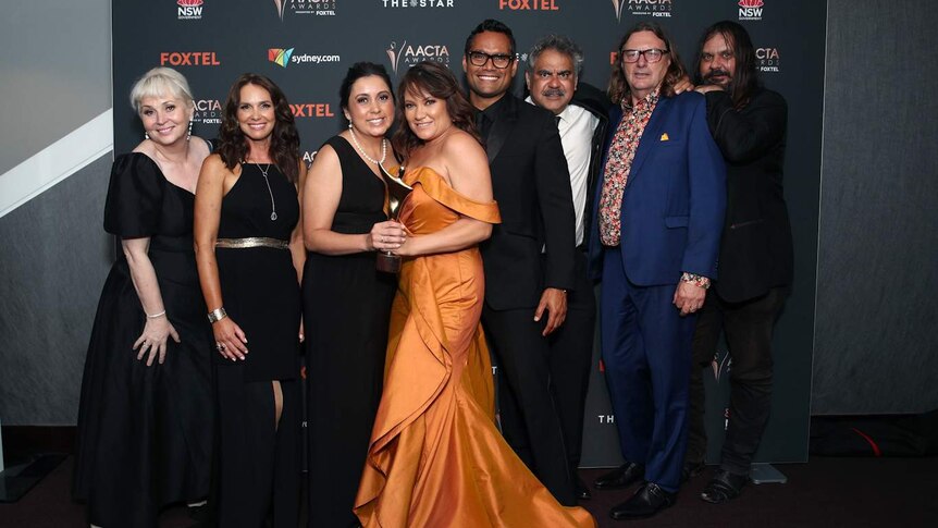 Line of eight people standing close with two women at centre smiling and holding AACTA Award trophy.