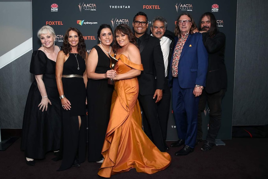 Line of eight people standing close with two women at centre smiling and holding AACTA Award trophy.