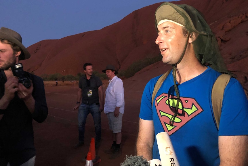 A white man wearing a hat with flaps and a Superman t-shirt is interviewed at the base of Uluru.