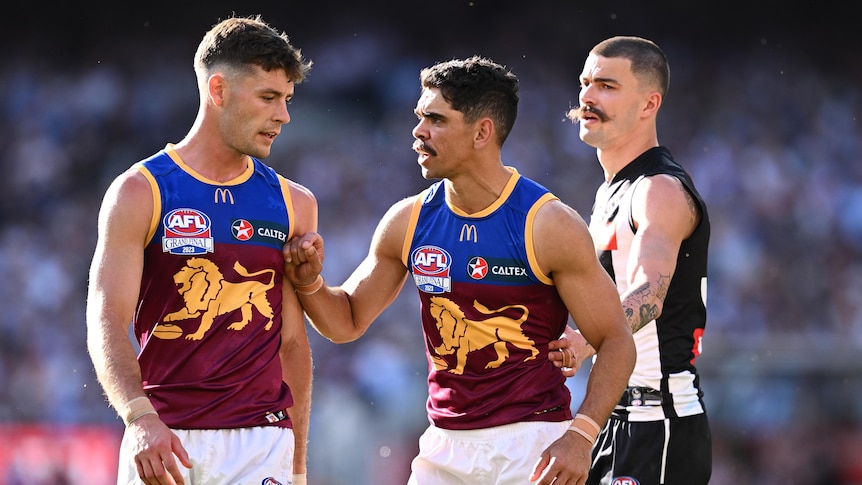 Two Brisbane Lions players stand together at the MCG, with one putting his hand up against his teammate as he speaks to him.