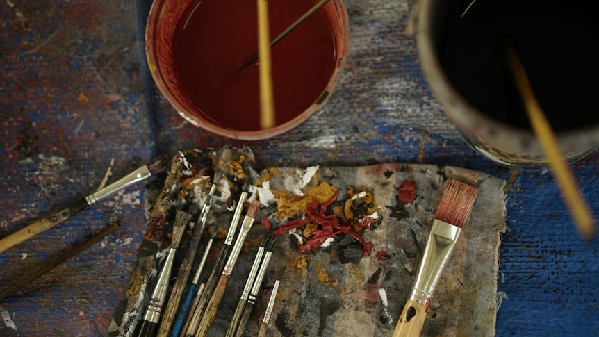 Used paint brushes sit on a piece of cloth surrounded by globs of paint and buckets of water