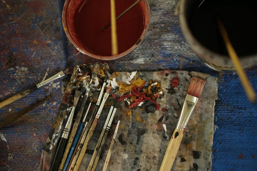 Used paint brushes sit on a piece of cloth surrounded by globs of paint and buckets of water