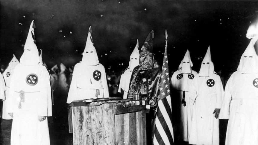 men stand around an alter wearing white robes and hooded masks.