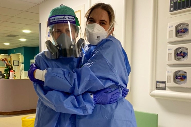 Two women in blue plastic protective clothes and wearing face masks hug in a hospital corridor