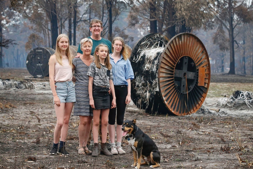 Cate Tregellas, her husband Mark and their three daughters stand with their dog in burnt bushland.