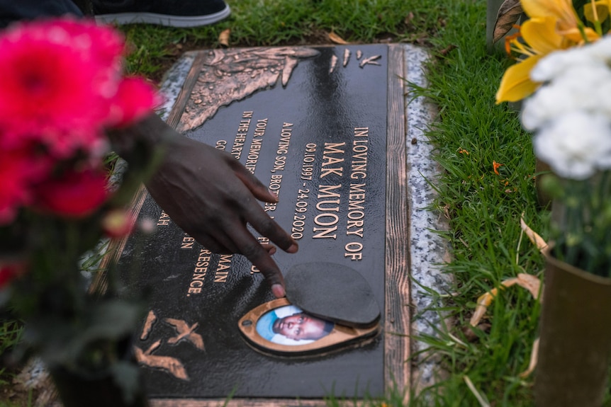 A hand touches the edge of a childhood portrait integrated into the plaque of a grave with the name "Mak Muon".