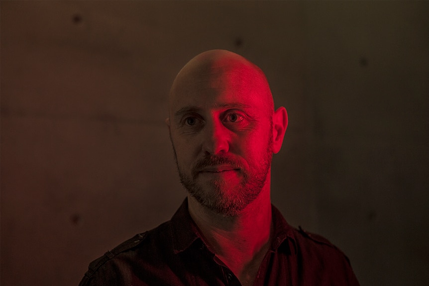 The composer stands against a bare, concrete wall, lit in dramatic red.