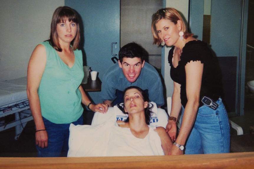 A frail woman in a hospital bed with husband and two sisters standing beside