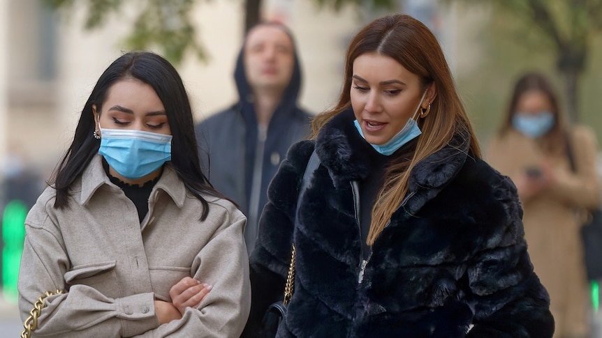 Young women, one wearing a mask, in an outdoor setting.