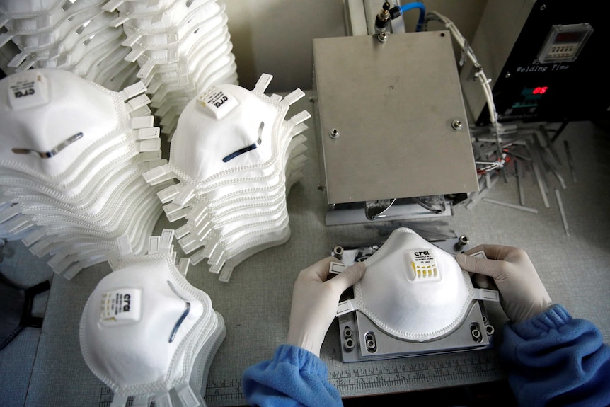 A bird's eye view shows dozens of white face masks stacked on to of each other next to a face mask-press with hands over it.