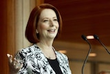 Julia Gillard stands at a lectern smiling and wearing a black and white patterned blazer over a black top.