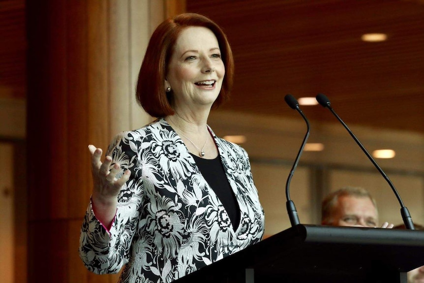 Julia Gillard stands at a lectern smiling and wearing a black and white patterned blazer over a black top.