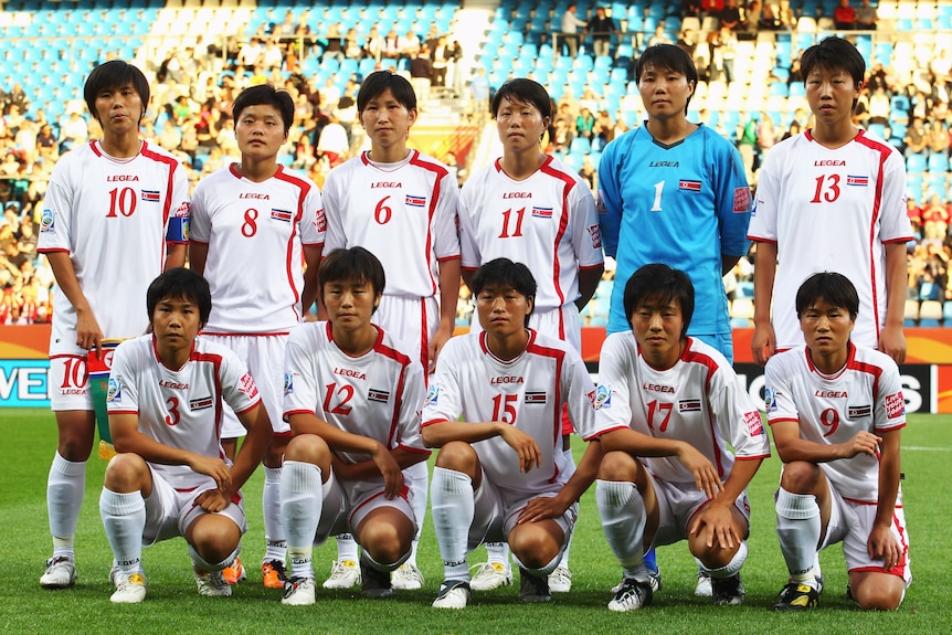 A women's soccer team wearing red and white pose for a photo with a crowd behind them before a game