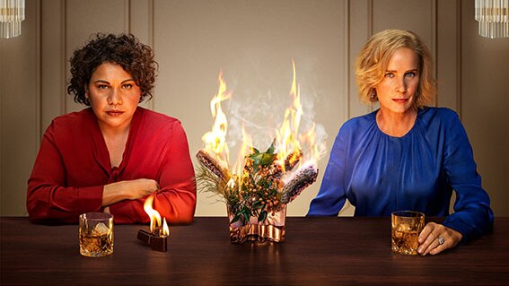 Deborah Mailman and Rachel Griffiths sit together at table with burning flowers in between them