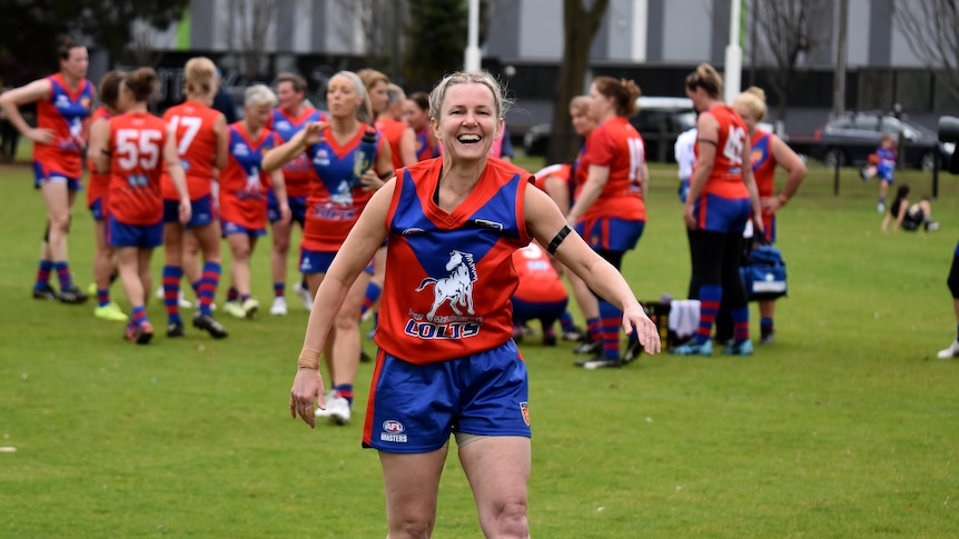 A woman in a blue and red Aussie Rules guernsey and shorts walks towards camera and smiles.