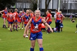 A woman in a blue and red Aussie Rules guernsey and shorts walks towards camera and smiles.