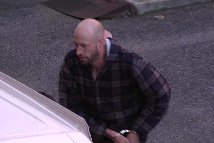A bald man with a beard wearing handcuffs and a plaid shirt