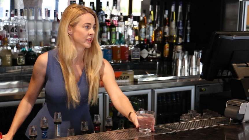 Ms Maddox stands behind the bar, placing a glass on the bar in front of her. Bottles of liquor are behind her.