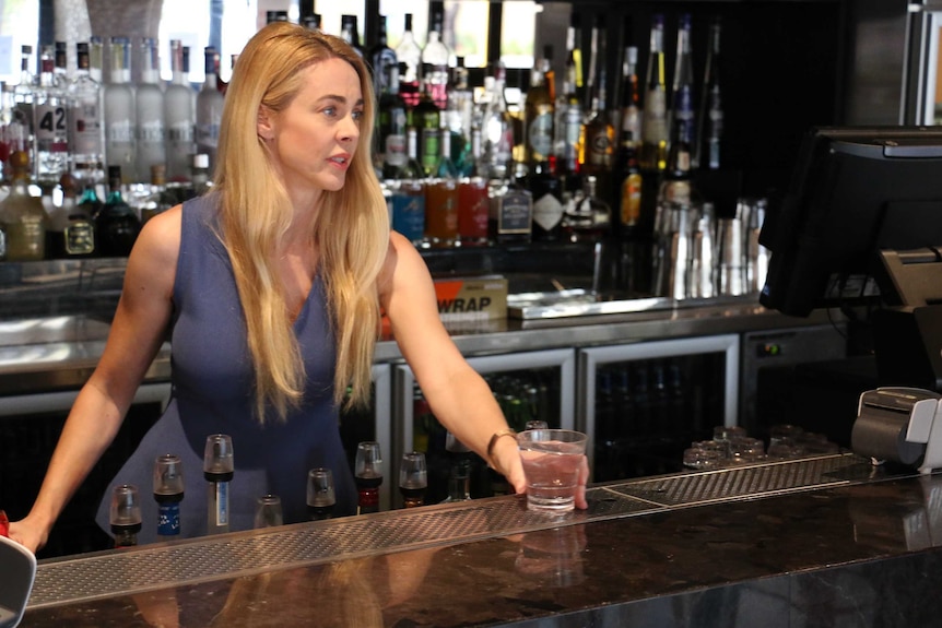 Ms Maddox stands behind the bar, placing a glass on the bar in front of her. Bottles of liquor are behind her.
