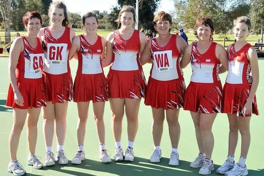 Seven women in red and white netball dresses pose for a photo on a netball court.