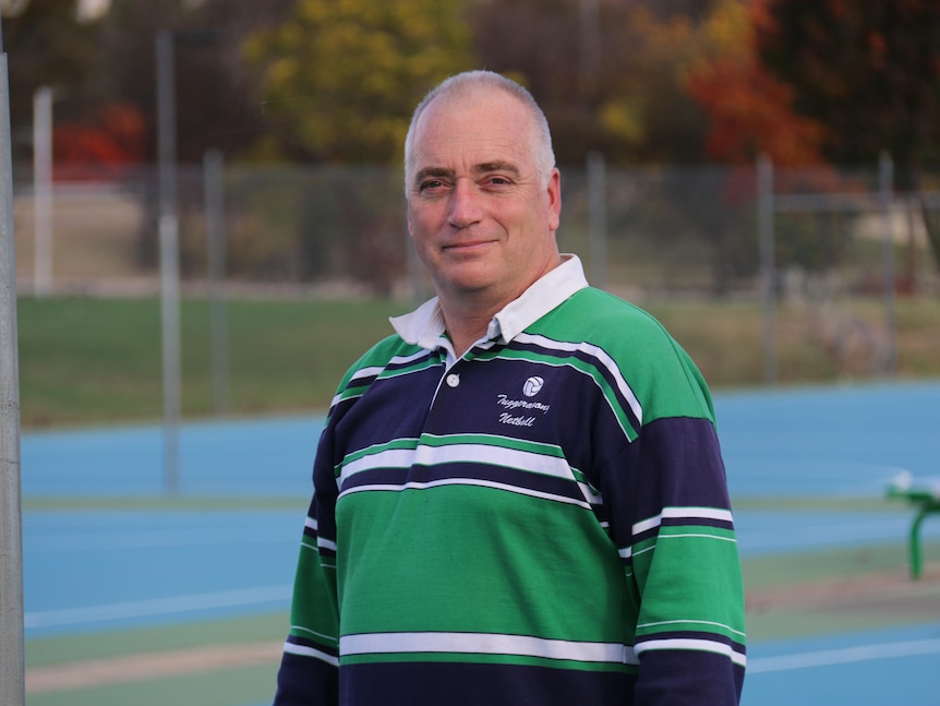 A man in a green jersey stands in front of a series of netball courts.