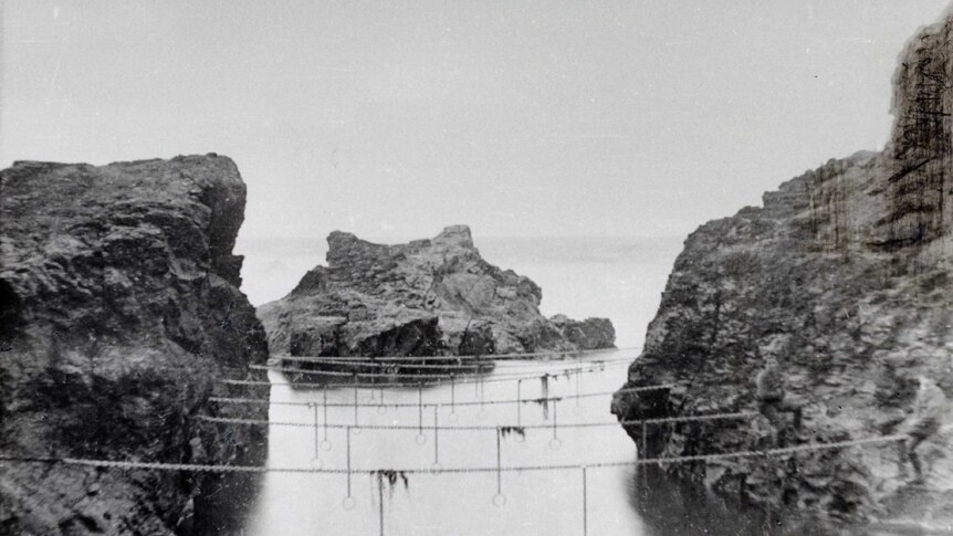 Black and white photo of ocean pool between cliffs. Metal rings hang from 7 horizontal chains