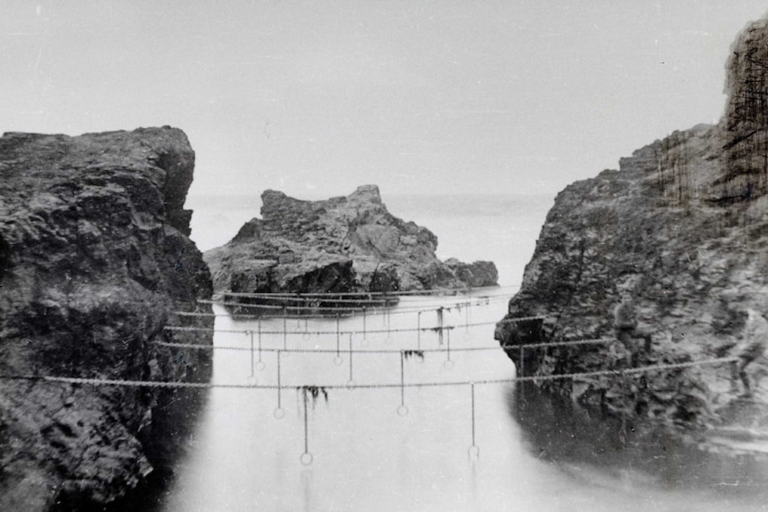 Black and white photo of ocean pool between cliffs. Metal rings hang from 7 horizontal chains