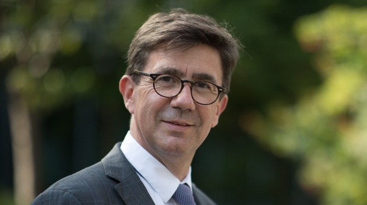 A man wearing glasses and a blue and grey suit smiles at the camera