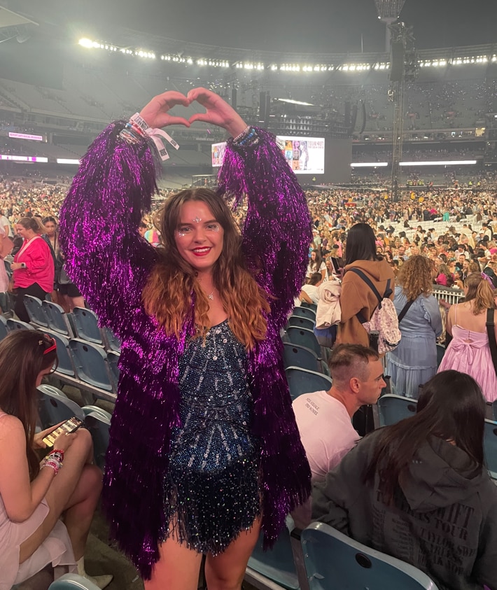 Una mujer joven, vestida con un deslumbrante leotardo azul medianoche y una chaqueta de oropel violeta, crea un corazón con sus manos.